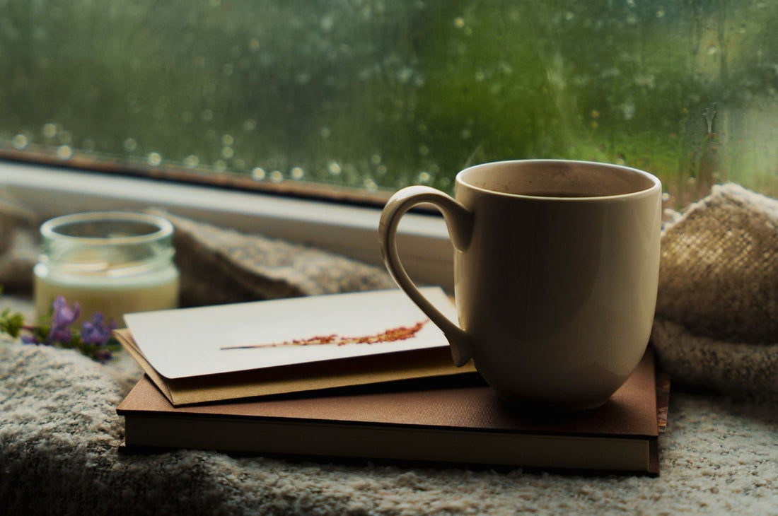 Cozy rainy day scene with a steaming mug, candle, and book by a rain-speckled window