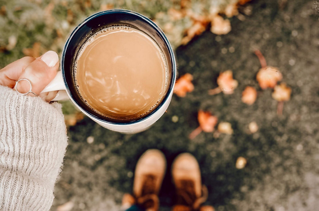 Hand holding a warm mug of coffee outdoors on a fall day, with autumn leaves and brown boots in the background.