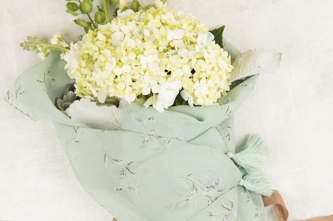 A white hydrangea bouquet wrapped in a pale green patterned scarf with mint tassels on a light background.
