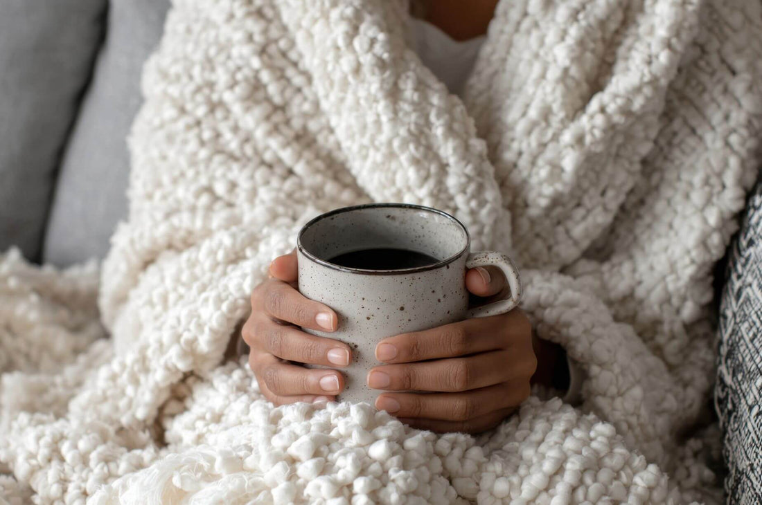 Close-up of a woman wrapped in a soft white blanket holding a speckled ceramic mug of tea, creating a cozy and calming winter moment.