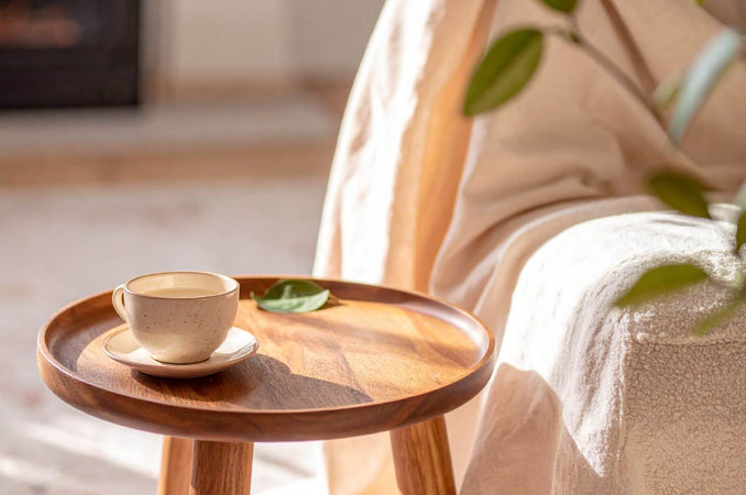 A ceramic mug on a wooden side table beside a cozy armchair with a soft knit throw, warm natural light, hygge home moment