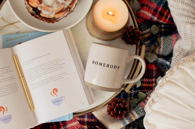 Cozy fall scene with a “Homebody” mug, lit candle, open book, plaid blanket, pinecones, and soft pillows.