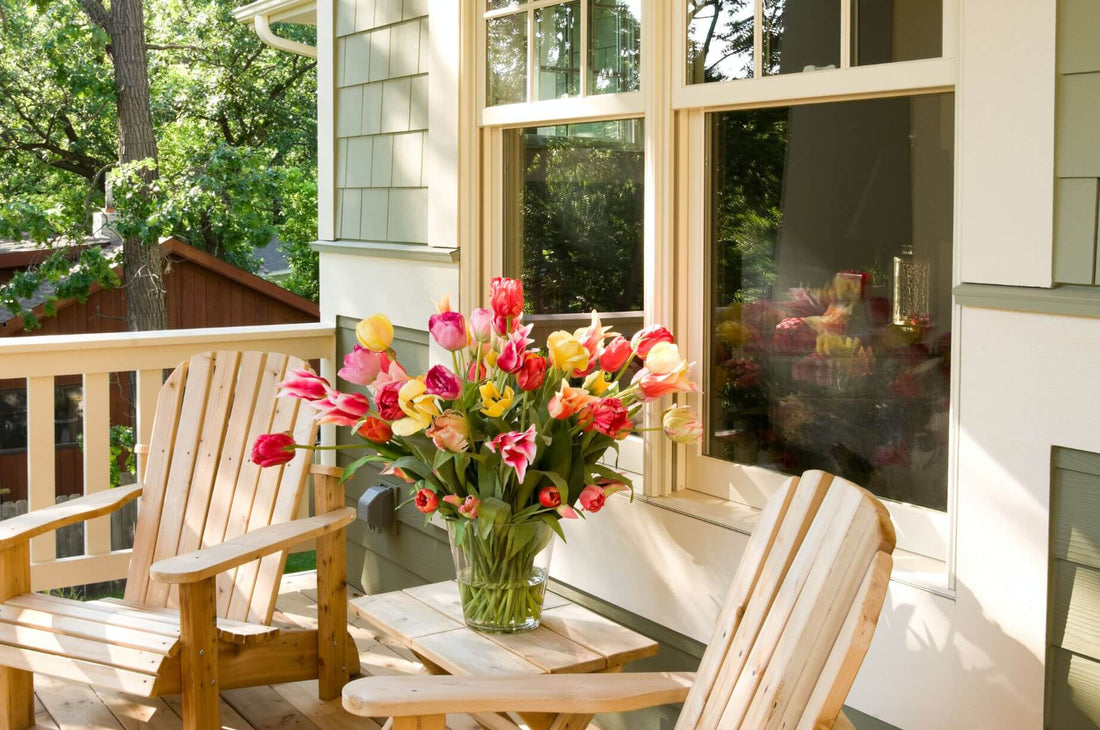 Colorful tulips in a glass vase on a sunlit porch table between two wooden Adirondack chairs.