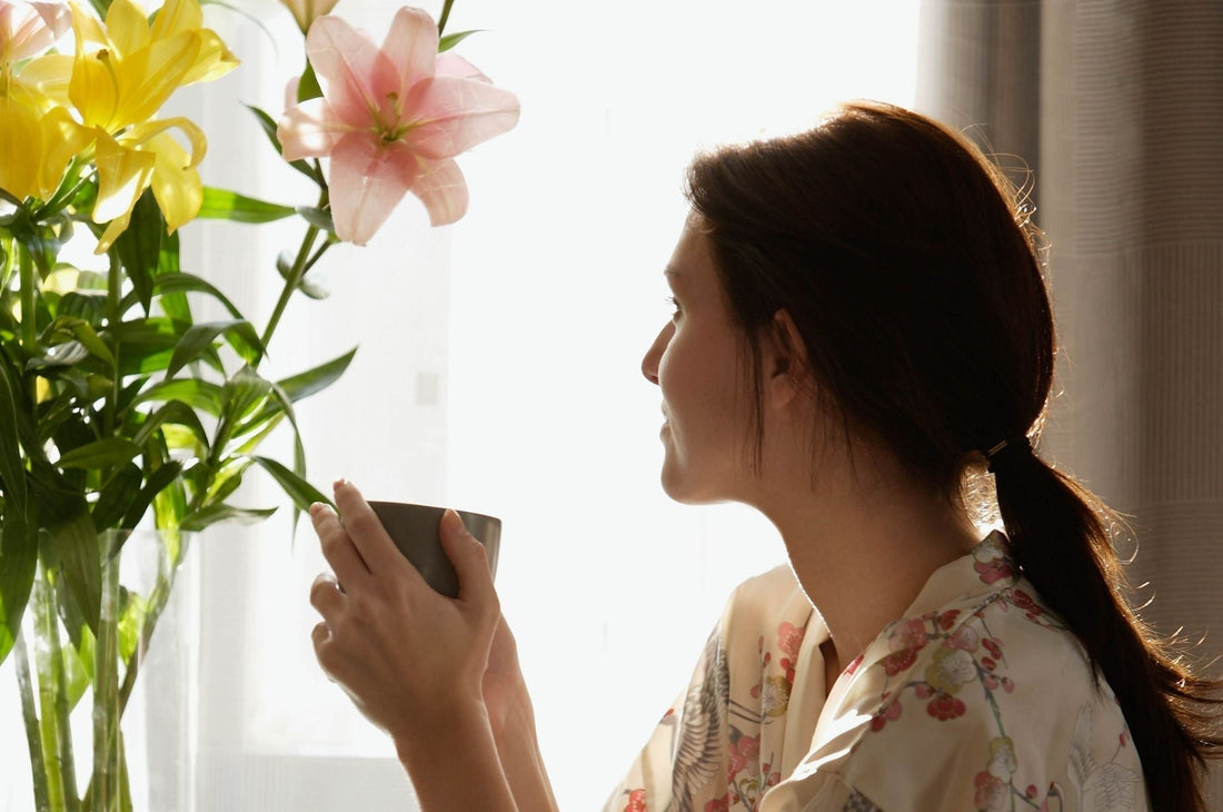 A woman in a floral robe sits by a bright window, holding a warm mug and gazing thoughtfully at a vase of fresh lilies, bathed in soft morning light.