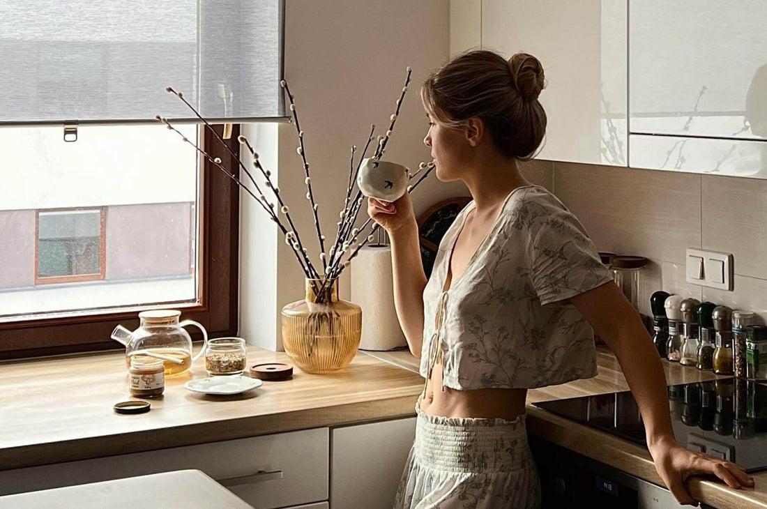 Woman standing by a sunlit kitchen window, enjoying a quiet drink and a peaceful moment at home.