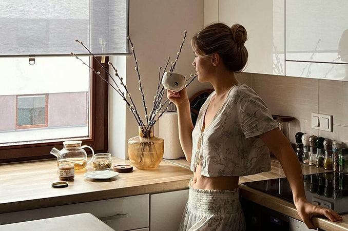 Woman standing by a sunlit kitchen window, enjoying a quiet drink and a peaceful moment at home.