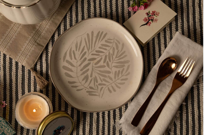 Warm, hygge-inspired table setting with a botanical ceramic plate, striped tablecloth, linen napkin, bronze flatware, lit candle, and small floral card.