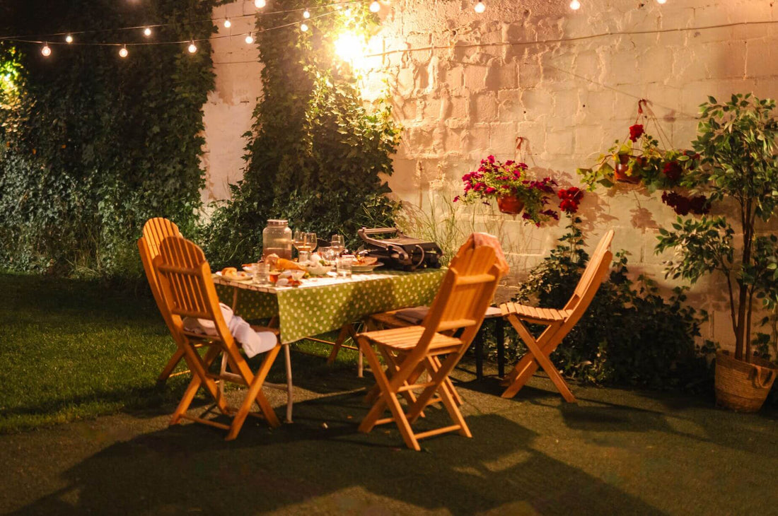 Outdoor late summer dinner setting with wooden chairs, string lights, flowers, and a table set in a cozy garden at dusk.