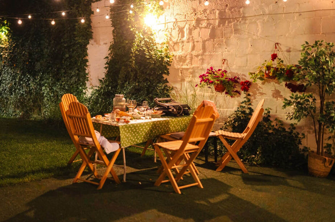 Outdoor late summer dinner setting with wooden chairs, string lights, flowers, and a table set in a cozy garden at dusk.