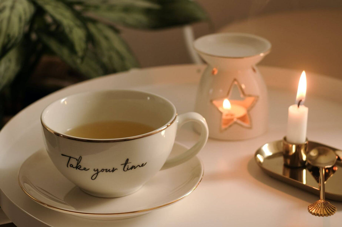 Cup of tea beside a glowing candle and ceramic tealight holder on a cozy table.