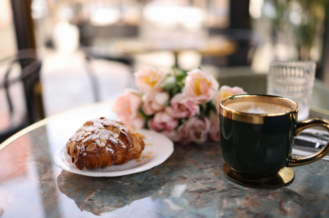A marble café table with a dark green and gold coffee cup, an almond croissant on a white plate, and a bouquet of pink roses in the background.