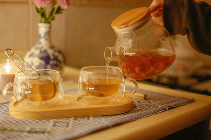 Tea being poured into a glass cup beside another cup, with a candle and flowers in the background.