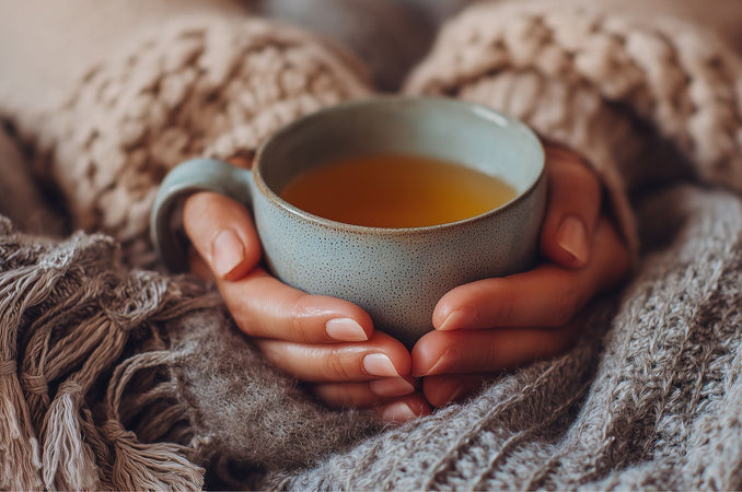 Hands holding a warm mug of tea while wrapped in cozy knit blankets and sweater.