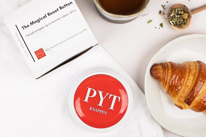 Red PYT Button on a white surface beside tea, a croissant, and its packaging.