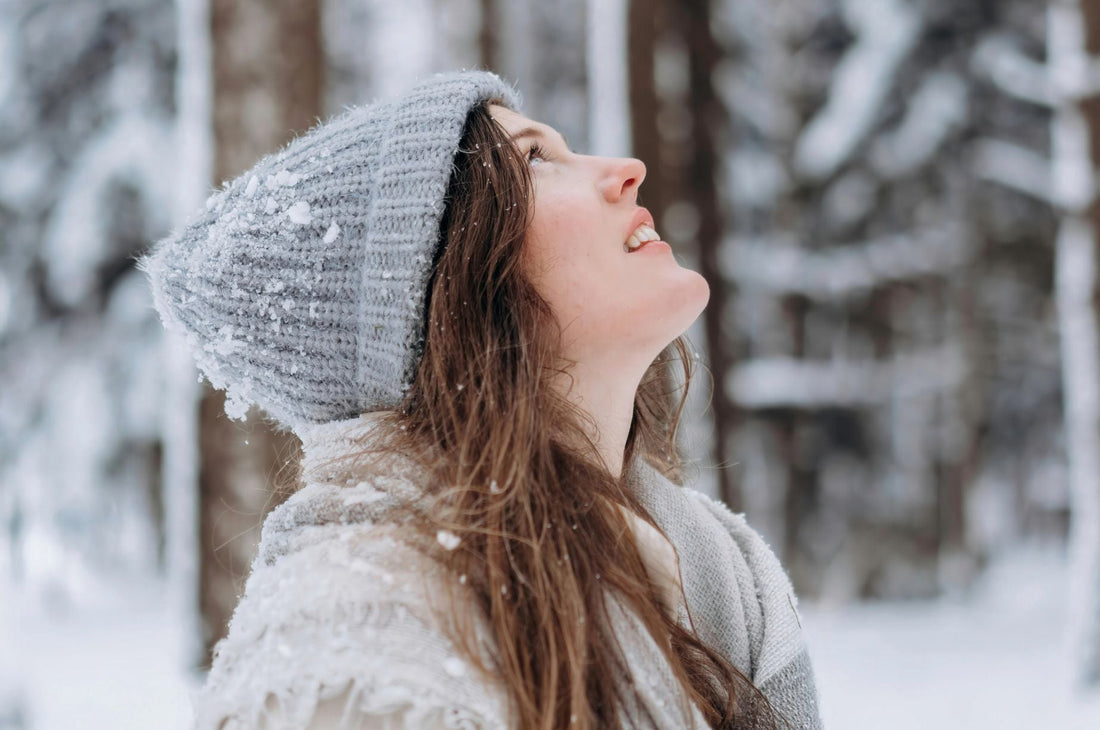 woman wearing a gray knit hat in a winter forest looking up
