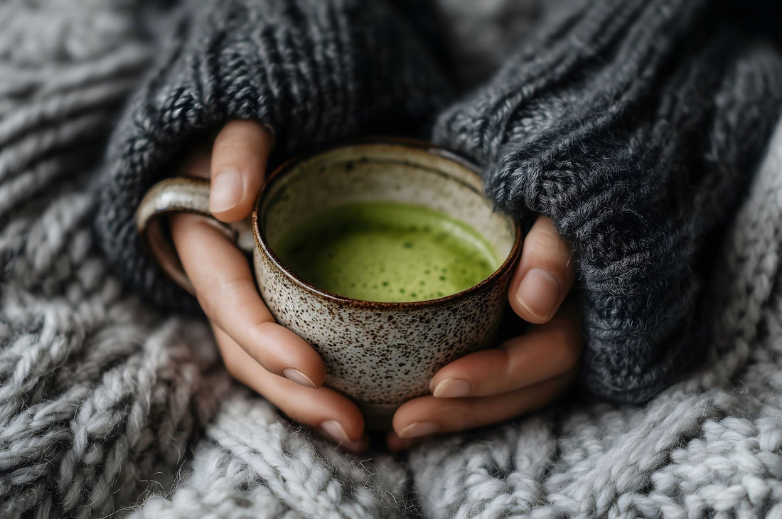 woman holding a mug of matcha between her hands laying on top of a knit throw.