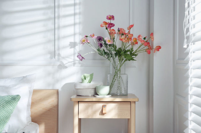 Wooden nightstand with spring flowers in a glass vase beside a neatly made bed with soft morning light filtering through window blinds.