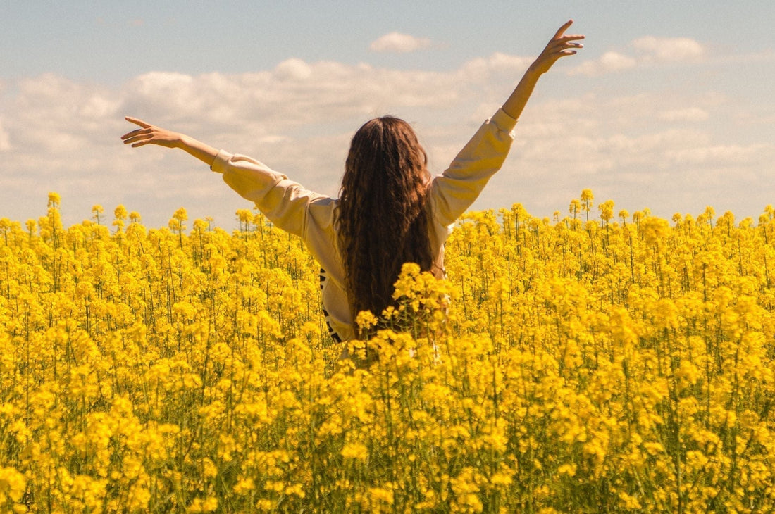 Woman standing in a sunny yellow flower field with arms raised, embracing summer sunshine and outdoor wellness.