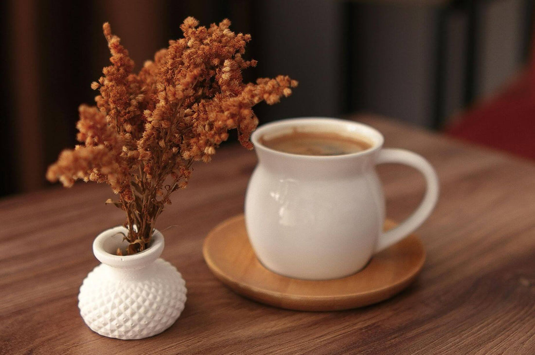 White ceramic mug of coffee on a wooden table beside a small vase of dried rust-colored flowers, creating a warm hygge-inspired self-care moment.