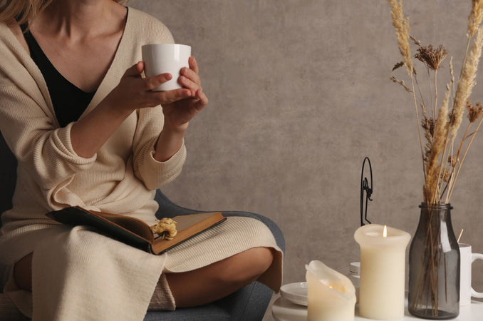 Woman in a cream cardigan holding a white mug, sitting cross-legged with an open book on her lap. A nearby table holds lit candles and dried grasses in a vase, creating a warm, calming atmosphere.