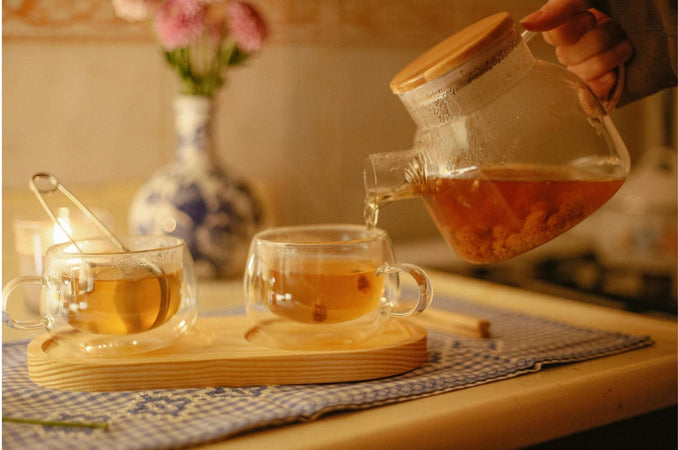 A hand pours hot tea from a glass teapot into a clear cup beside another cup on a wooden tray, lit by warm candlelight with a small vase of flowers softly blurred in the background.
