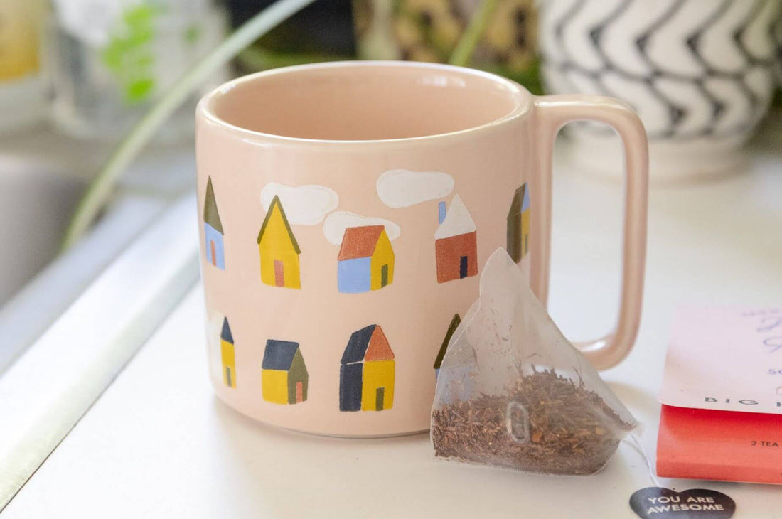 Pink ceramic mug decorated with colorful little houses beside a tea bag, creating a relaxing, cozy tea moment.