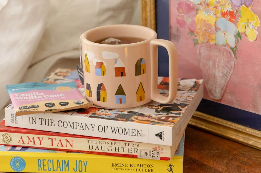 Cozy pink mug with house illustrations on a stack of books and floral art in the background.