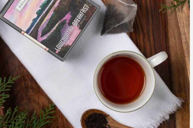 Overhead view of a cup of black tea on a white cloth napkin beside a “Lowcountry Breakfast” tea box and loose-leaf tea on a wooden table.