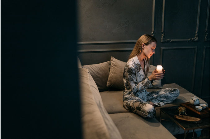 A woman in patterned pajamas sits cross-legged on a cushioned sofa in a dim, cozy room, gently holding a lit candle beside a tea set, creating a calm hygge evening ritual atmosphere.