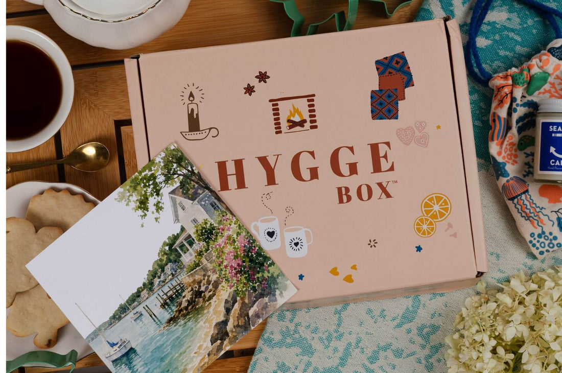 July Hygge Box on a cozy tabletop with tea, cookies, a postcard, flowers, and summer-inspired accessories arranged around the Hygge Box.