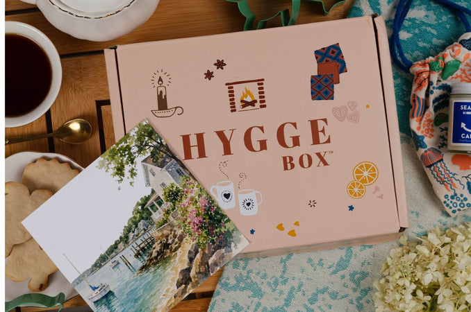 July Hygge Box on a cozy tabletop with tea, cookies, a postcard, flowers, and summer-inspired accessories arranged around the Hygge Box.