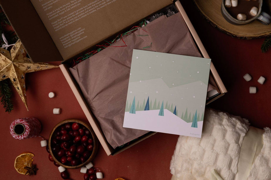 Open Hygge Box Subscription Box with colorful tissue paper and illustrated winter card, surrounded by cranberries, marshmallows, dried orange slices, and a soft ivory blanket.