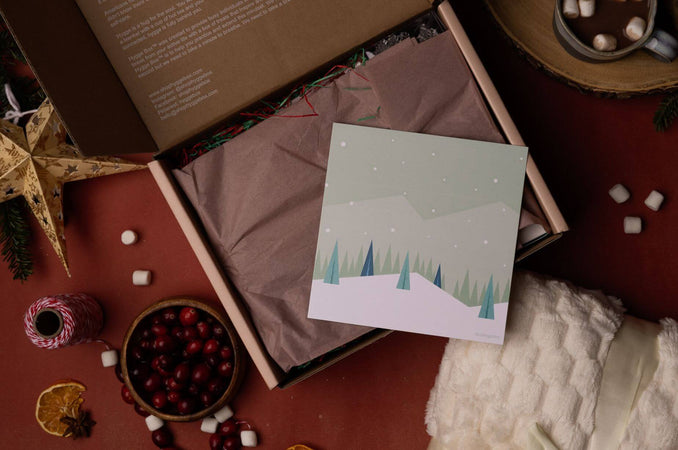 Open Hygge Box Subscription Box with colorful tissue paper and illustrated winter card, surrounded by cranberries, marshmallows, dried orange slices, and a soft ivory blanket.