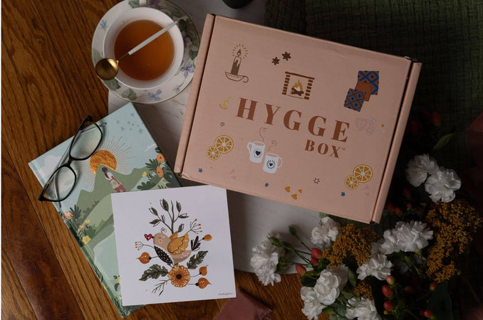 Hygge Box surrounded by a cup of tea, reading glasses, the Nature Almanac, the Awaken Gently card, and fresh white flowers on a wooden table