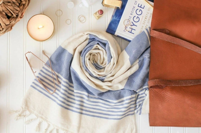 Blue and cream striped Turkish towel beside a candle, hygge book, glasses, and brown leather bag.