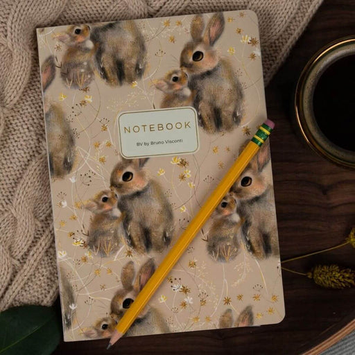 A notebook with a beige cover decorated with watercolor-style rabbits and small gold floral details, placed on a dark wooden surface with a yellow pencil resting diagonally across it. A knitted beige textile and part of a cup are also visible around the notebook.