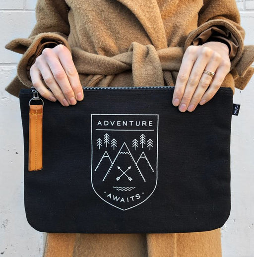 Woman wearing a tan wool coat holding a black canvas folio with a leather zipper pull and white graphic design featuring mountains, trees, crossed oars, and the words 'Adventure Awaits.