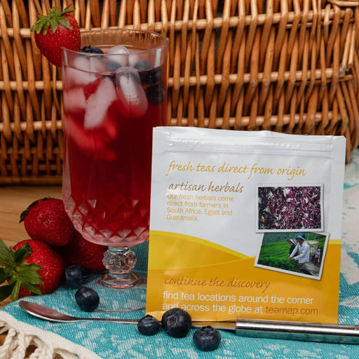 A stand-up pouch of tea placed on a turquoise towel beside a glass of berry iced tea, with strawberries, blueberries, a spoon, and a wicker basket in the background. The pouch reads 'fresh teas direct from origin' and 'artisan herbals'.