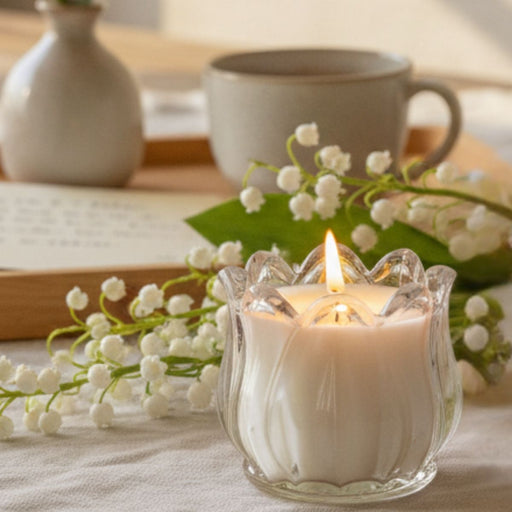 Candle in a tulip shaped decorative holder with lily of the valley flowers and a cup in the background.