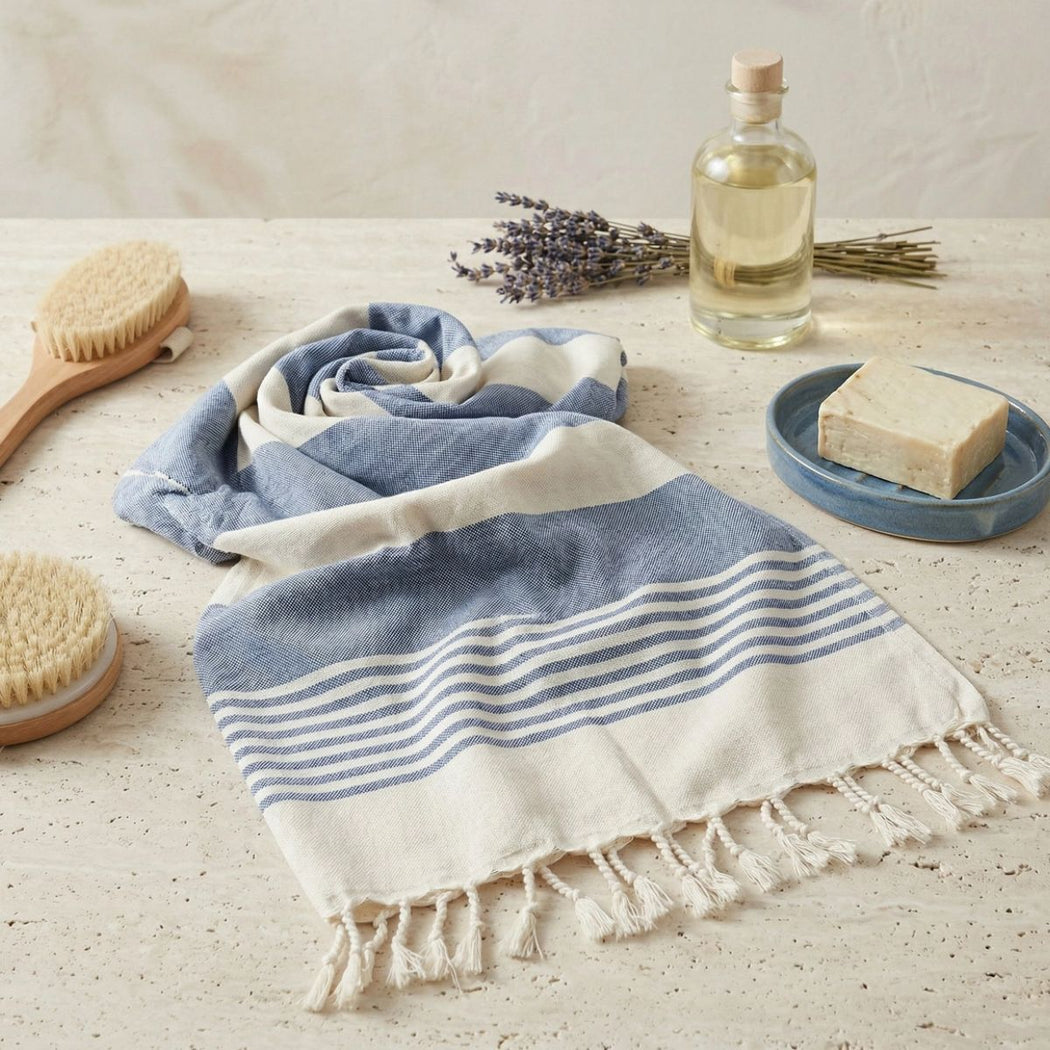 A folded cotton Turkish towel with blue and white stripes and fringed edges is styled on a light stone surface with spa props including a wooden brush, lavender, a bottle of oil, and a bar of soap.
