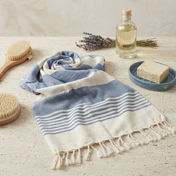 A folded cotton Turkish towel with blue and white stripes and fringed edges is styled on a light stone surface with spa props including a wooden brush, lavender, a bottle of oil, and a bar of soap.