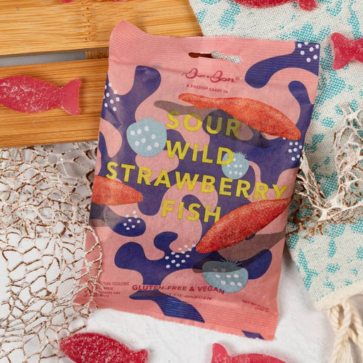 A bag of BonBon Sour Wild Strawberry Fish candy is displayed on a white textured surface with nautical props. The candy fish pieces are scattered around the bag.