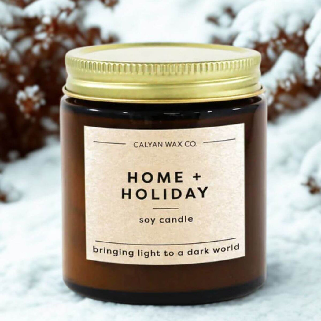 Candle with 'Home + Holiday' label in front of snow-covered bushes