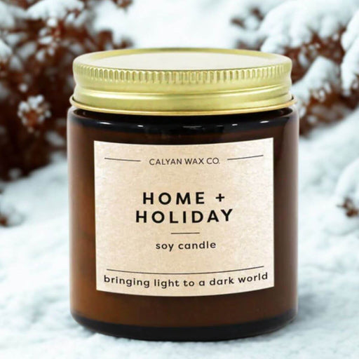 Candle with 'Home + Holiday' label in front of snow-covered bushes