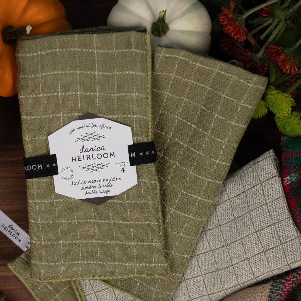Two woven kitchen towels with a windowpane plaid pattern in sage green and light beige are arranged on a dark rustic surface with decorative pumpkins and flowers.