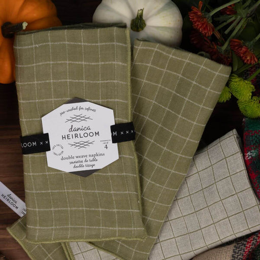 Two woven kitchen towels with a windowpane plaid pattern in sage green and light beige are arranged on a dark rustic surface with decorative pumpkins and flowers.