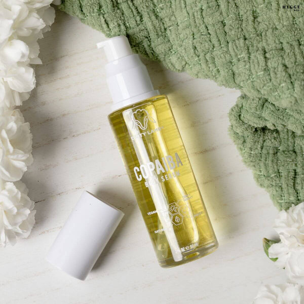 Bottle of Copaiba oil serum with a white pump on a light wooden surface with green towel and flowers.
