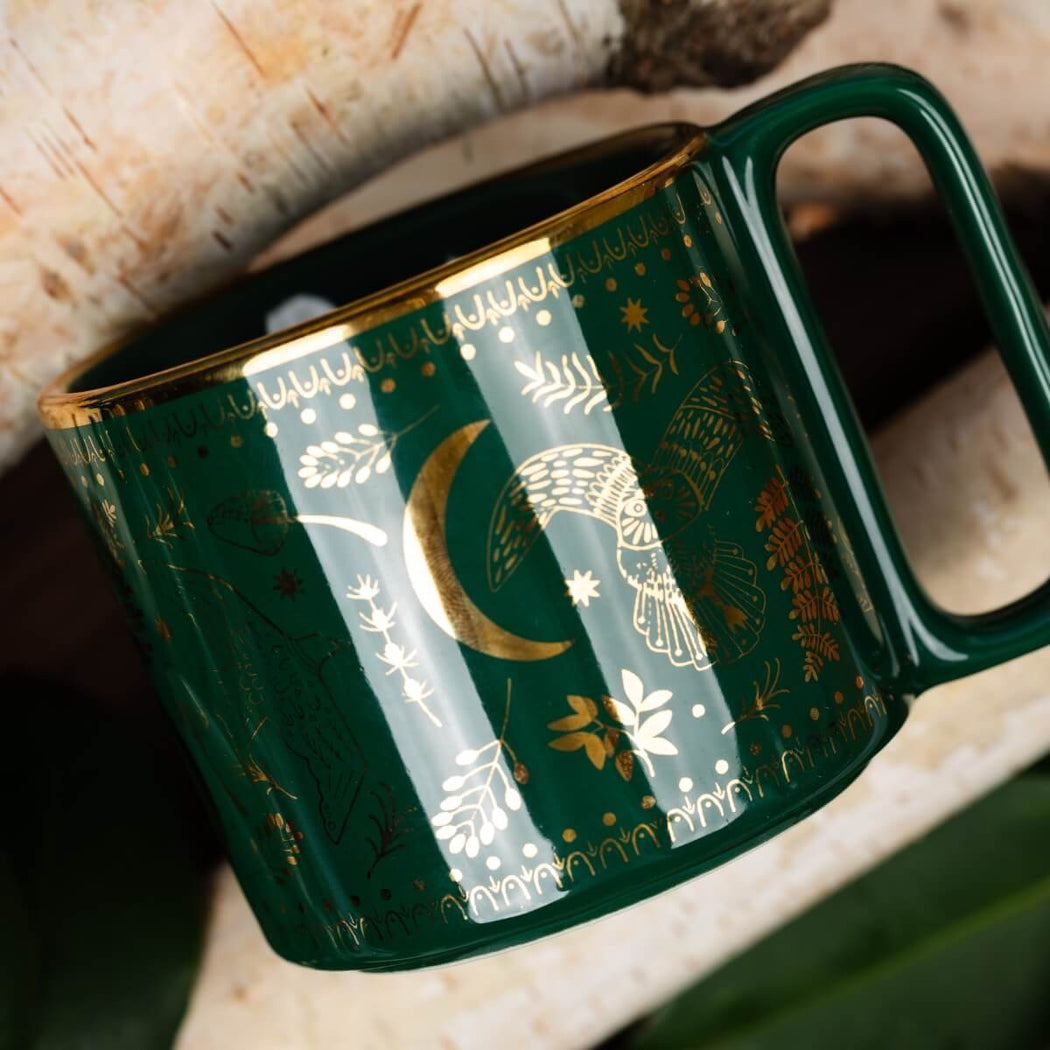 A dark green ceramic mug with gold celestial illustrations, including a crescent moon, owl, stars, and floral motifs, resting on a rustic wood surface.