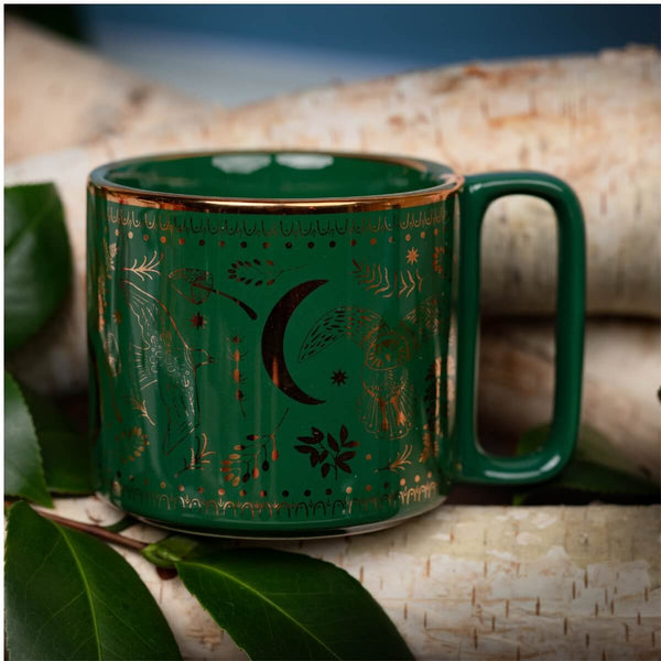 A dark green ceramic mug with gold celestial and nature illustrations, including a crescent moon, owl, stars, and leaves, resting on a birch log with green leaves in the foreground.