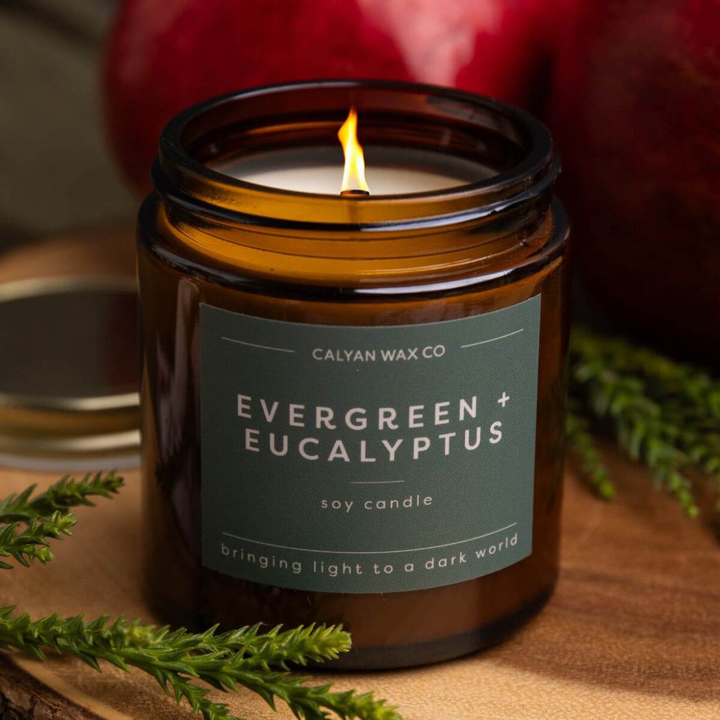 Candle in a jar labeled 'Evergreen + Eucalyptus' from Calyan Wax Co, with greenery around on a wooden surface.